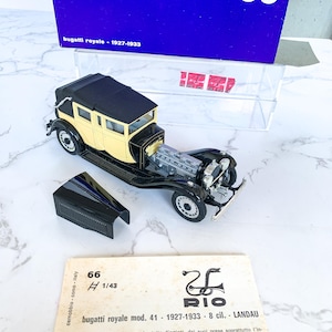May include: A black and cream 1/43 scale model of a Bugatti Royale Landau, a luxury car from the 1920s and 1930s. The model is displayed on a white surface with a blue box and a white card with the Rio logo and text about the car.