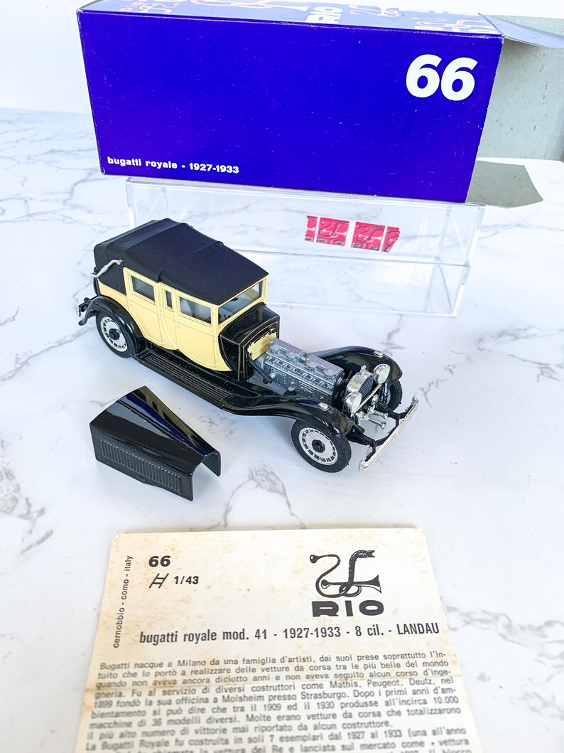 May include: A black and cream 1/43 scale model of a Bugatti Royale Landau, a luxury car from the 1920s and 1930s. The model is displayed on a white surface with a blue box and a white card with the Rio logo and text about the car.