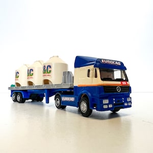 May include: A blue and white toy truck with a white trailer carrying three white cylindrical containers. The truck has the word "EUROCAB" printed on the side and the number "1850" on the front. The trailer has the word "EUROLAD" printed on the side.