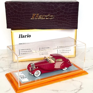 Könnte beinhalten: Ein burgunderfarbenes Modellauto in einer transparenten Vitrine mit Holzsockel. Das Auto hat Weißwandreifen und einen cremefarbenen Innenraum. Die Vitrine steht auf einer Marmoroberfläche. Auf der Box steht das Wort "Ilario" in goldfarbenen Buchstaben.