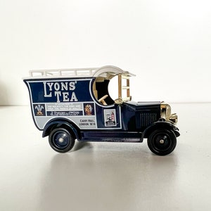 Puede incluir: Un camión de reparto de juguete azul con techo blanco y un letrero blanco que dice "Lyons Tea" en letras doradas. El camión tiene una parrilla dorada y ruedas doradas. El letrero también dice "His Majesty the King and HRH The Prince of Wales" y "J. Lyons & Co. Ltd. Cadby Hall, London W14."