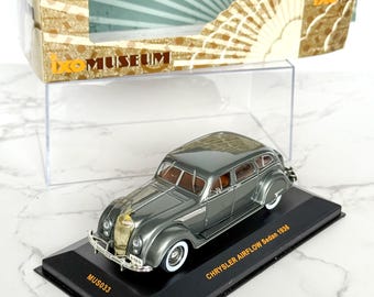 May include: A diecast model of a 1936 Chrysler Airflow Sedan in a metallic gray finish. The model sits on a black base with the text "CHRYSLER AIRFLOW Sedan 1936". The model is displayed in a clear plastic case with a decorative box.