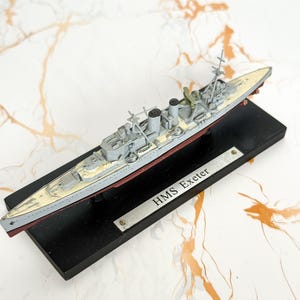 May include: A scale model of the HMS Exeter warship featuring a grey hull, a cream-colored deck, and a dark red lower section. The model is mounted on a rectangular black display base with a silver metal plaque engraved with the text HMS Exeter.