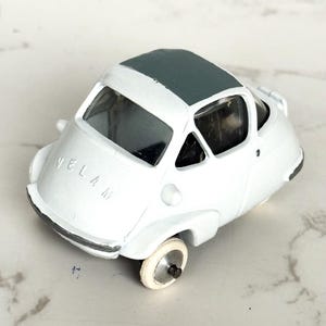 May include: A white toy car with a dark gray roof. The car has the word "VELAM" printed on the front. The wheels are white with silver rims. The car is a miniature replica of a vintage vehicle.