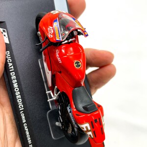 Może przedstawiać: Model motocykla Ducati Desmosedici w kolorze czerwonym z numerem 55 i nazwiskiem "Loris Capirossi" na boku. Motocykl stoi na czarnym stojaku wystawowym z napisem "DUCATI DESMOSEDICI Loris Capirossi 2004".
