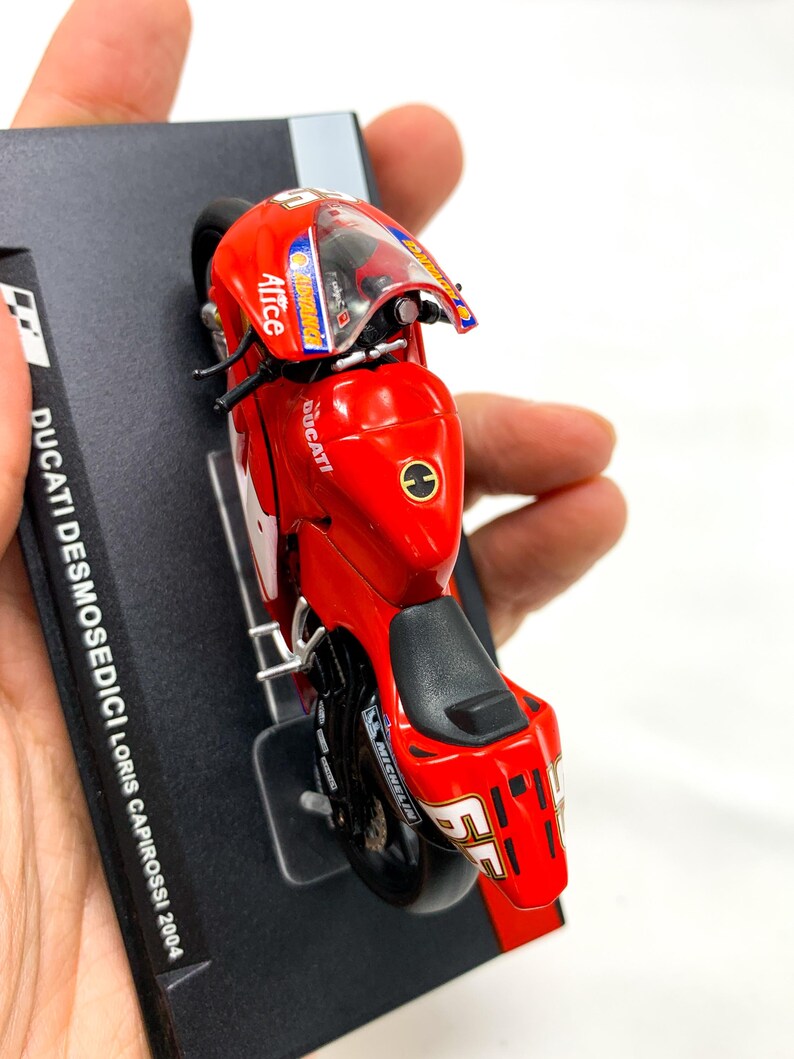 Może przedstawiać: Model motocykla Ducati Desmosedici w kolorze czerwonym z numerem 55 i nazwiskiem "Loris Capirossi" na boku. Motocykl stoi na czarnym stojaku wystawowym z napisem "DUCATI DESMOSEDICI Loris Capirossi 2004".
