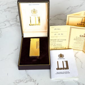May include: A gold Dunhill lighter in a brown box with gold lettering. The lighter has a textured surface. Included are warranty cards and a booklet. The box is lined with cream-colored material. Made in Switzerland.