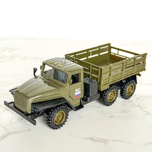 May include: Miniature olive green toy truck with a cargo bed. The model truck has six wheels, a detailed cab, and a black front bumper. The truck has the letters "BC" on the side. A replica.