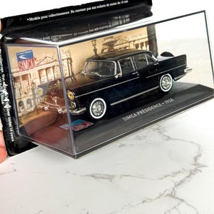 May include: A black Simca Présidence 1958 model car in a clear display case. The model car has white-walled tires and chrome details. The display case has a black base and a printed background with a city scene. The text on the car reads "SIMCA PRÉSIDENCE 1958".
