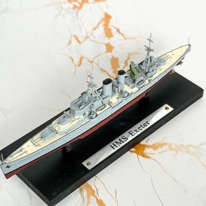 May include: A scale model of the HMS Exeter warship featuring a grey hull, a cream-colored deck, and black smokestacks. The ship is mounted on a rectangular black base with a silver metal plaque engraved with the text HMS Exeter.