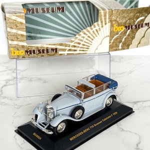 Könnte beinhalten: Hellblaues Modellauto Mercedes-Benz 770 Grosser Cabriolet F 1930 auf schwarzem Sockel. Das Modell hat ein blaues Cabrioverdeck und eine braune Innenausstattung. Die Box trägt die Aufschrift "ixo MUSEUM". Das Modell ist ca. 18 cm lang.