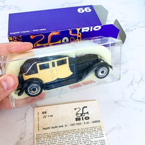 May include: A black and cream 1/43 scale model of a Bugatti Royale Landau, a luxury car from the 1920s and 1930s. The model is displayed in a clear plastic case with a blue and white box.