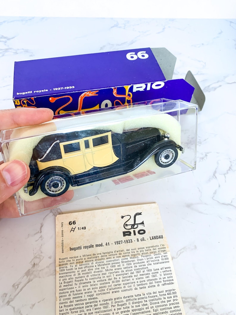 May include: A black and cream 1/43 scale model of a Bugatti Royale Landau, a luxury car from the 1920s and 1930s. The model is displayed in a clear plastic case with a blue and white box.