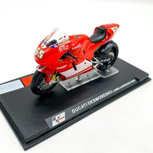 Peut inclure: Modèle de moto Ducati Desmosedici rouge et blanc avec le numéro 65 et le mot "Alice" sur le côté. Le modèle est sur un socle d'exposition noir avec le texte "DUCATI DESMOSEDICI LORIS CAPROSSI 2004".