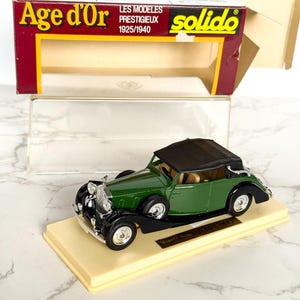 Könnte beinhalten: Ein grünes Modellauto mit schwarzem Cabrioverdeck, präsentiert auf einem beigen Ständer. Das Modell ist ein Oldtimer aus der "Age d'Or"-Kollektion, mit der Originalverpackung im Hintergrund.