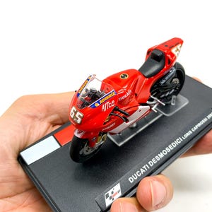 Może przedstawiać: Model motocykla Ducati Desmosedici w kolorze czerwonym i białym z numerem 65 na boku. Model znajduje się na czarnej podstawie wystawowej z nadrukiem "DUCATI DESMOSEDICI - Loris Capirossi 2003".