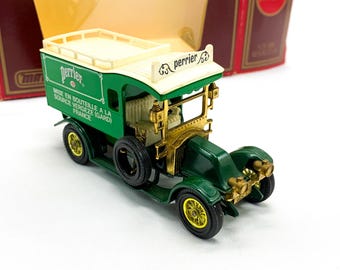May include: A green toy delivery truck with a white roof and a Perrier logo. The truck has gold accents and a white sign that reads "Perrier" in cursive. The truck is on a white background.