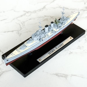 May include: A model of the HMS Exeter warship, painted in shades of gray, white, and red, mounted on a black base. The model features detailed structures, including turrets and masts. A small metal plate on the base reads "HMS Exeter".