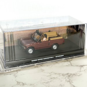 Puede incluir: Modelo de un Range Rover descapotable burdeos en una vitrina transparente. El modelo tiene un interior beige y ruedas plateadas. El texto "RANGE ROVER CONVERTIBLE • OCTOPUSSY" está impreso en la base.