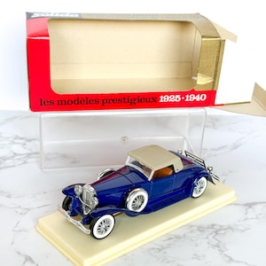 May include: A die-cast model of a vintage blue convertible car with a tan roof and white-walled tires. The car sits on a cream-colored base, with the box in the background displaying the text "les modèles prestigieux 1925-1940".