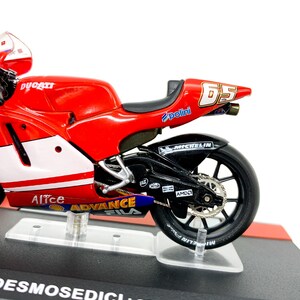 Może przedstawiać: Model motocykla Ducati w kolorze czerwonym i białym z numerem 65 na boku. Motocykl stoi na czarnej podstawie wystawowej z napisem "Desmosedici Loris Capirossi 2004".