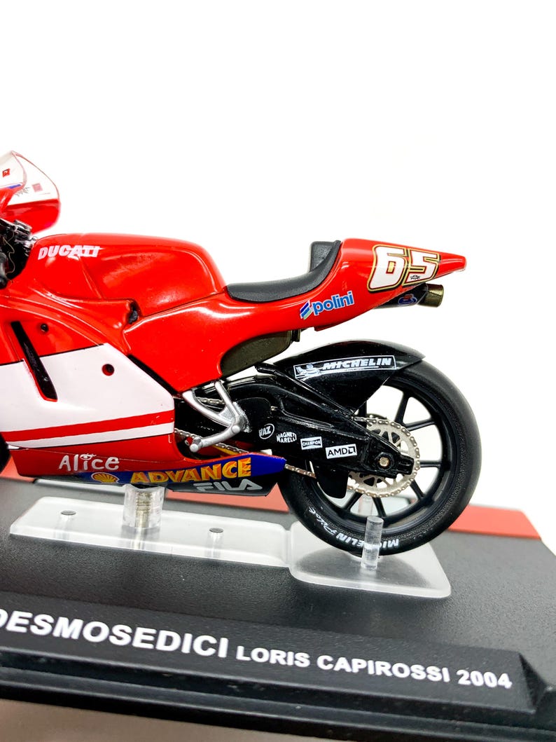 Może przedstawiać: Model motocykla Ducati w kolorze czerwonym i białym z numerem 65 na boku. Motocykl stoi na czarnej podstawie wystawowej z napisem "Desmosedici Loris Capirossi 2004".