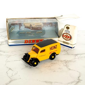 Könnte beinhalten: Ein gelb-schwarzes Heinz-Lieferwagen-Spielzeug mit der Aufschrift "HEINZ 57 VARIETIES". Das Spielzeug ist ein Dinky-Modell mit schwarzen Rädern und Dach. Die Originalverpackung ist im Hintergrund zu sehen. Das Modell ist ca. 10 cm lang.