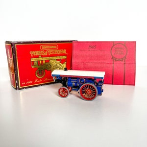 Op de afbeelding: Een blauw en rood speelgoed stoommachine model met een wit baldakijn. De machine staat op een wit oppervlak. De doos voor het speelgoed is op de achtergrond. De doos is rood en bruin met de woorden "Matchbox Models of Yesteryear" en "Y19 1905 Fowler Thomas Engine".