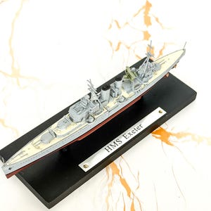 May include: A scale model of the HMS Exeter warship featuring a light gray hull, a cream-colored deck, and black smokestacks. The model is mounted on a rectangular black base with a silver-toned metal plaque engraved with the text HMS Exeter.