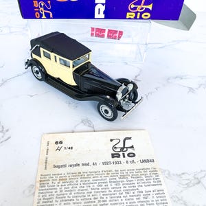 May include: A black and cream 1/43 scale model of a Bugatti Royale Landau, a luxury car from the 1920s. The model is displayed on a white surface with a box in the background. The box has a red and blue design with the word "RIO" printed on it.