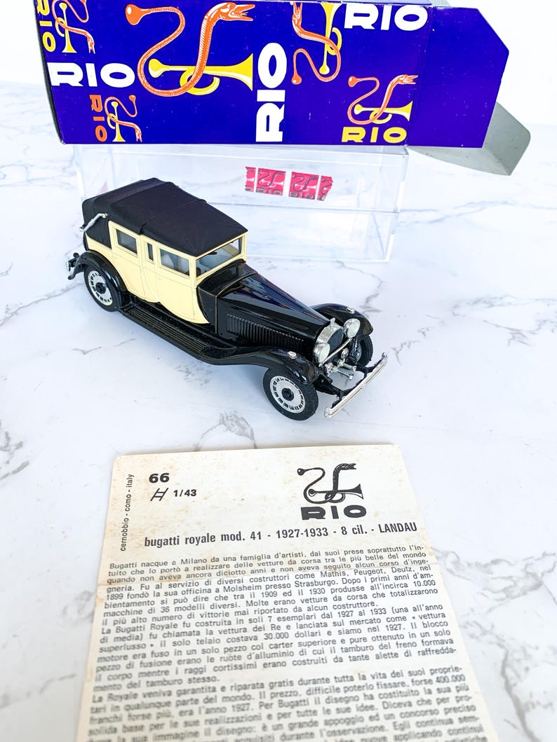 May include: A black and cream 1/43 scale model of a Bugatti Royale Landau, a luxury car from the 1920s. The model is displayed on a white surface with a box in the background. The box has a red and blue design with the word "RIO" printed on it.