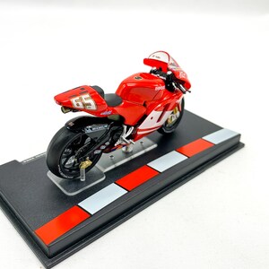 Może przedstawiać: Model motocykla Ducati w kolorze czerwonym i białym z numerem 69 na boku. Motocykl stoi na czarnym stojaku z czerwonymi i białymi szachownicami.