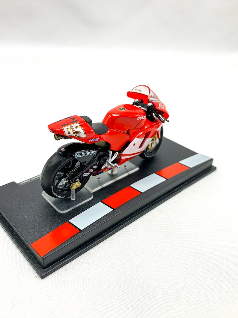 Może przedstawiać: Model motocykla Ducati w kolorze czerwonym i białym z numerem 69 na boku. Motocykl stoi na czarnym stojaku z czerwonymi i białymi szachownicami.