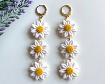 May include: A pair of gold dangle earrings with three white daisy flowers on each earring. The daisies have yellow centers.