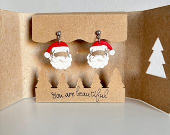 May include: Santa Claus earrings with red hats and white beards are displayed on a brown cardboard stand. The stand has a decorative tree-shaped edge and the handwritten text "You are beautiful". The earrings are presented in a folded cardboard gift box.