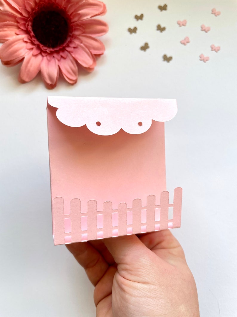 May include: A pink paper card with a scalloped edge and a cut-out design of a picket fence. The card has two holes punched at the top.