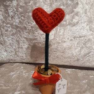 May include: A small terracotta pot with a red crocheted heart on a green stem. The pot is filled with small stones and has a red ribbon bow. A white tag with the text "Just for You" is attached to the pot.