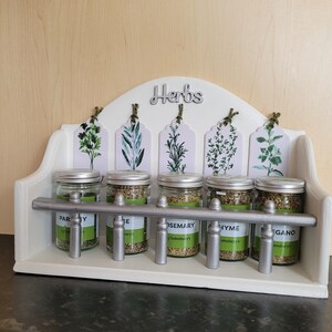 May include: White wooden spice rack with the word "Herbs" on the top. The rack has five glass jars with silver lids and labels that say "Parsley", "Sage", "Rosemary", "Thyme", and "Oregano".