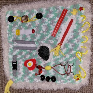 May include: A green and white sensory play mat with various textures and objects, including a zipper, a brush, a buckle, a red and yellow egg, and a yellow string with beads.