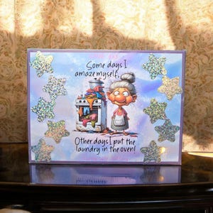 May include: A humorous greeting card with a cartoon illustration of a woman with a surprised expression. The card features the text "Some days I amaze myself, Other days I put the laundry in the oven!" and is decorated with sparkly stars.