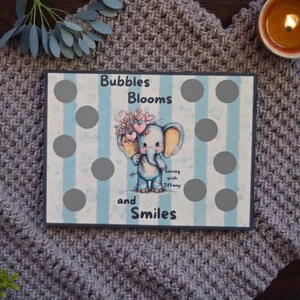 May include: A rectangular wooden board with a blue and white striped background, featuring a cartoon elephant holding flowers and hearts. The text "Bubbles Blooms and Smiles" is displayed. There are gray circles on the board.