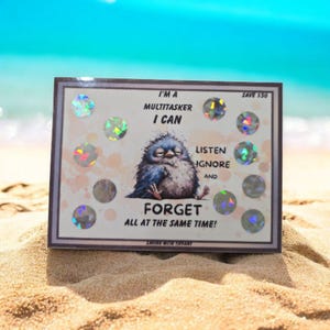 May include: A framed print with the text "I'M A MULTITASKER I CAN LISTEN IGNORE AND FORGET ALL AT THE SAME TIME!" features a cartoon bird. The print has holographic circles and is set on a sandy beach.