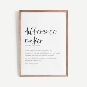 May include: A framed print with the words "difference maker" in a stylish script font. The definition of "difference maker" is included below the title in a smaller, more readable font. The frame is a light brown color.