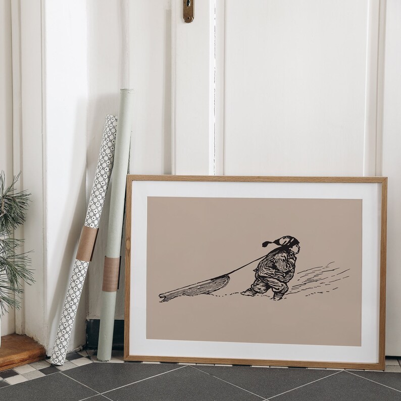 May include: A framed print featuring a black and white illustration of a child pulling a sled through the snow. The artwork has a light brown background and is encased in a wooden frame. Two rolls of decorative paper lean against the wall.