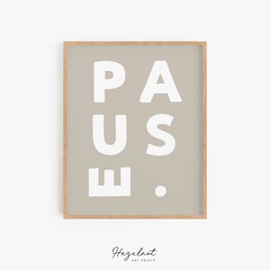 May include: A framed art print featuring the word "PAUSE" in bold white letters against a neutral backdrop. The artwork is presented in a light wooden frame, with the artist's name, "Hazelnut Art Prints," displayed at the bottom. A minimalist design.