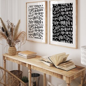 Puede incluir: Dos impresiones art&iacute;sticas abstractas enmarcadas con patrones de pinceladas en blanco y negro cuelgan sobre una consola de madera. La mesa exhibe libros, una cesta de mimbre y objetos decorativos. Un jarr&oacute;n con flores secas a&ntilde;ade un elemento natural a la habitaci&oacute;n.