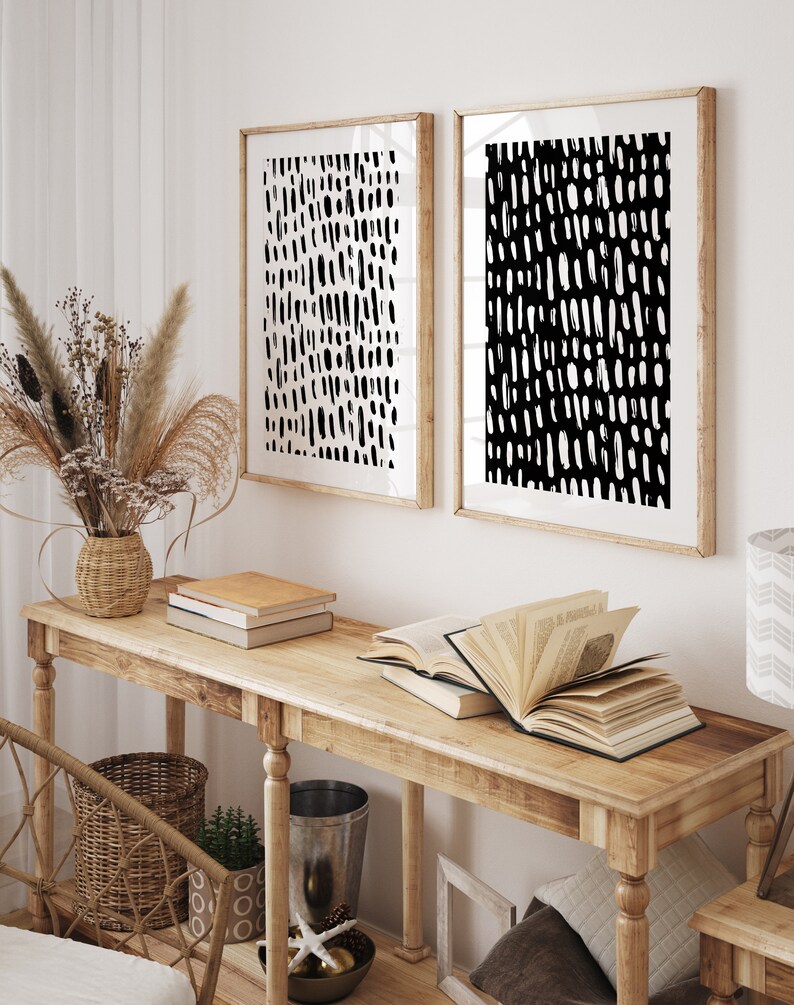 Puede incluir: Dos impresiones art&iacute;sticas abstractas enmarcadas con patrones de pinceladas en blanco y negro cuelgan sobre una consola de madera. La mesa exhibe libros, una cesta de mimbre y objetos decorativos. Un jarr&oacute;n con flores secas a&ntilde;ade un elemento natural a la habitaci&oacute;n.
