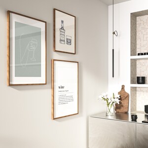 Op de afbeelding: Drie ingelijste kunstdrukken met houten frames hangen aan een lichtgrijze muur. E&eacute;n print toont een lijntekening van een hand die een wijnglas vasthoudt. Een andere toont een whiskyfles en een glas. De derde print heeft de definitie van "wijn".
