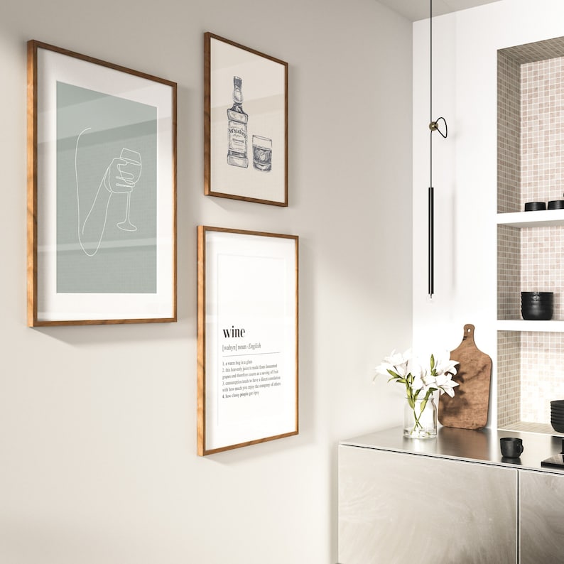 Op de afbeelding: Drie ingelijste kunstdrukken met houten frames hangen aan een lichtgrijze muur. E&eacute;n print toont een lijntekening van een hand die een wijnglas vasthoudt. Een andere toont een whiskyfles en een glas. De derde print heeft de definitie van "wijn".