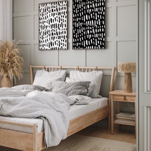 Puede incluir: Dos impresiones art&iacute;sticas abstractas enmarcadas con patrones de pinceladas en blanco y negro cuelgan sobre una cama con marco de madera clara y ropa de cama blanca. Una mesita de noche de madera con una l&aacute;mpara y libros se encuentra junto a la cama.